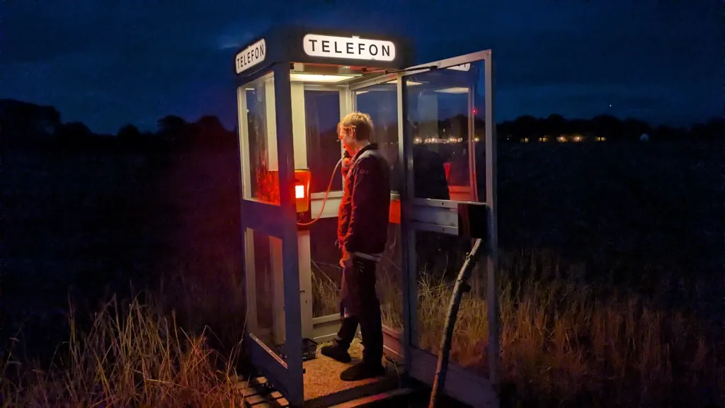 A visitor standing inside the booth on a call at night, lit red from the phone